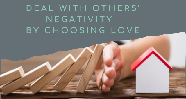 Green text on a grey background says "Deal with others negativity by choosing love". Beneath is a photo of wooden blocks falling and stopped by a hand before they hit a toy house