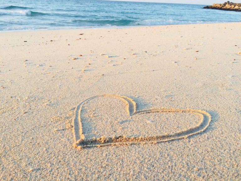 On a sandy beach, a heart has been carved into the sand. The sea is in the distance.