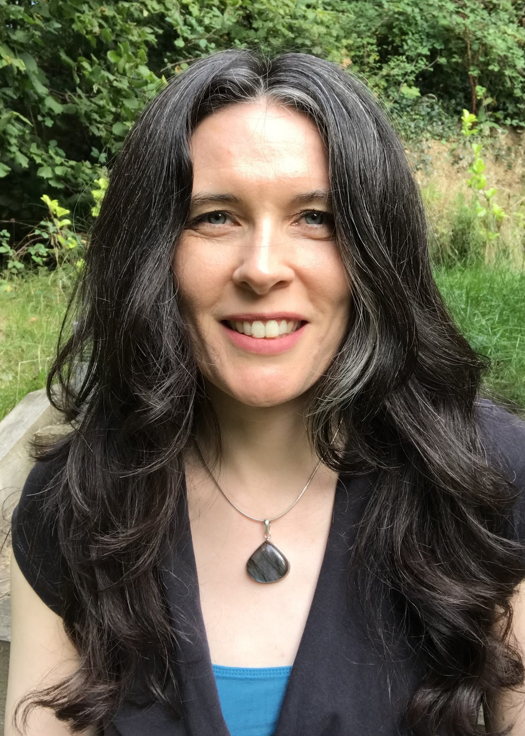 Headshot of Jacqui McGinn who has long, wavy, dark hair and white skin. She's outside with greenery behind her and wearing a labradorite crystal around her neck.