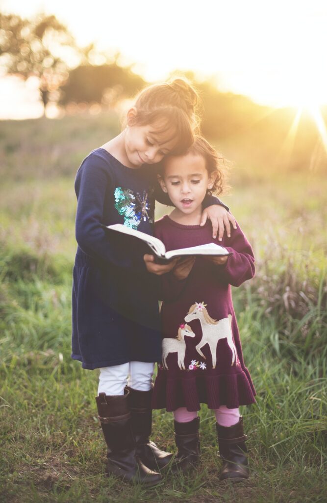 1 older girl about 7 years old is wearing a navy dress and putting her arm around a younger girl wearing a burgundy dress. The older girl is holding a book and they they are both standing up reading it. They're outside in a field with the light of the setting sun in the top right corner of the photo.