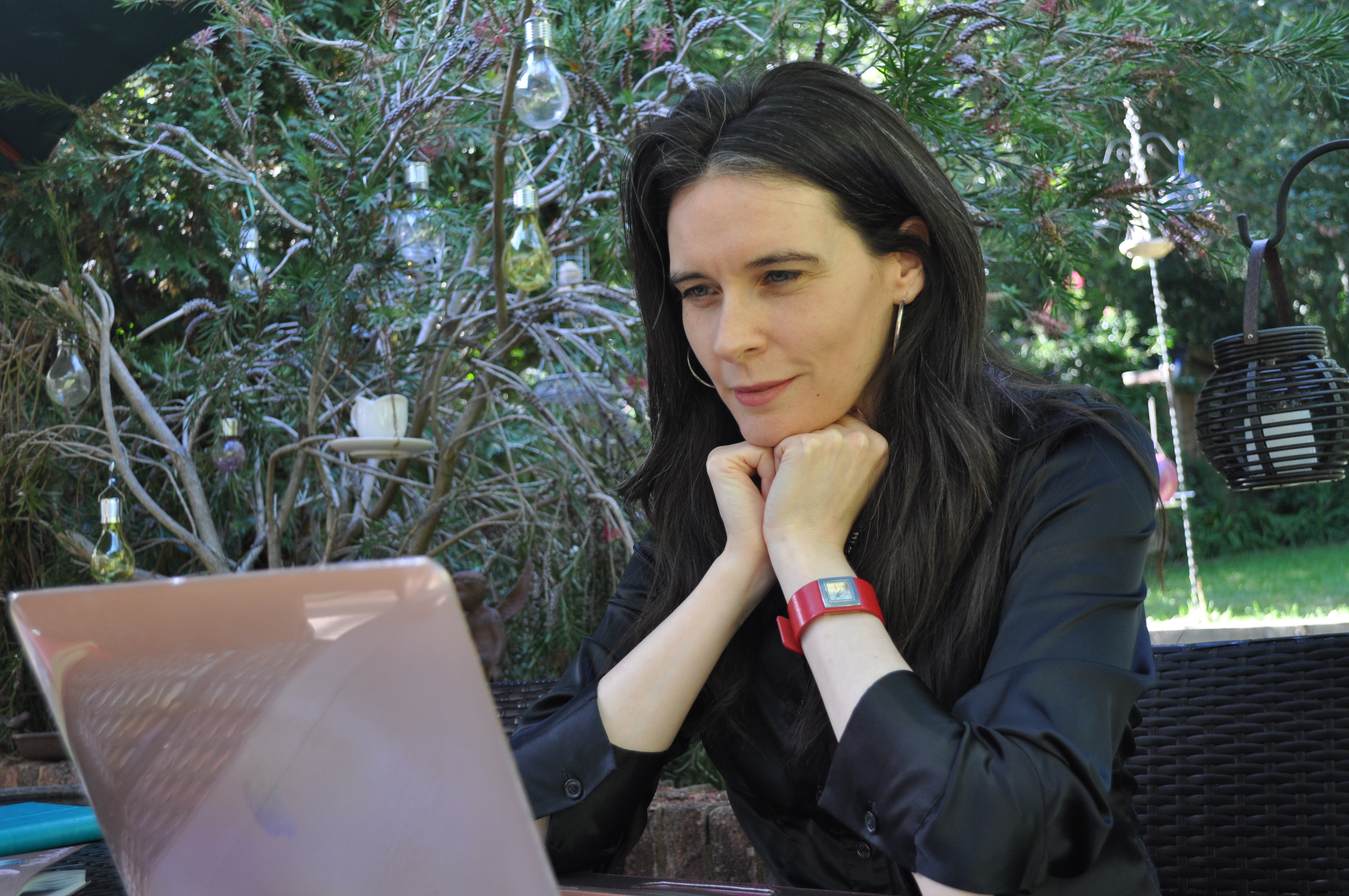 Jacqui McGinn (dark-haired white woman wearing a black blouse) looks down at her MacBook with her chin resting on her hands. A tree and garden are behind her.