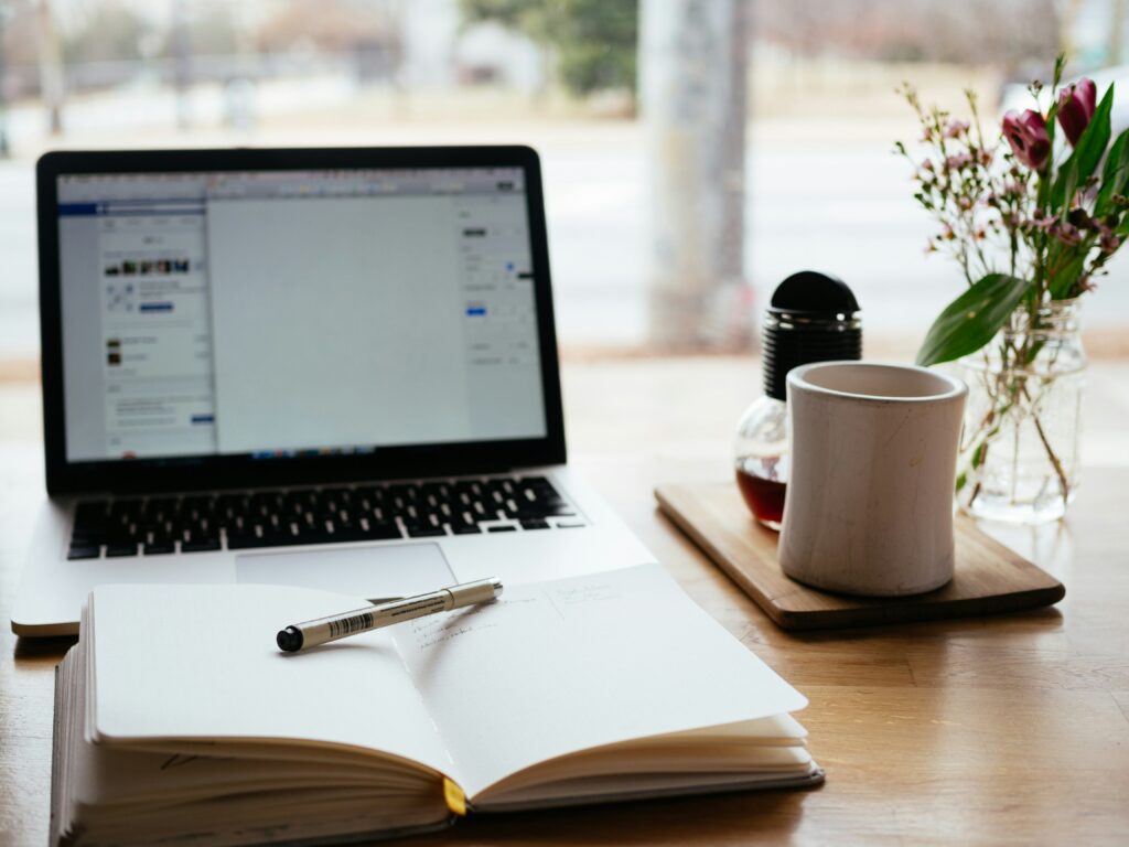 A computer, cup of coffee, journal and pen on a desk in front of a window.